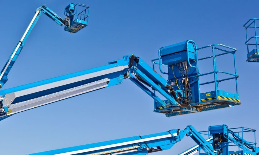 Blue boom-type elevating work platforms (cherry pickers) with articulated booms and personnel baskets raised against a clear sky, illustrating equipment used for working at heights.
