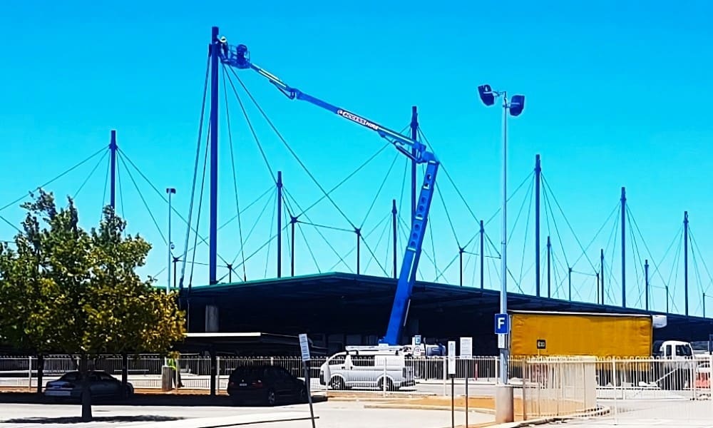 Blue boom-type elevating work platform (articulated boom lift) extended to lift a worker or equipment high above ground at a work site against a clear blue sky.
